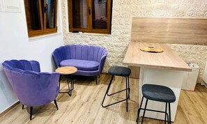 A small room with a wooden table, two purple chairs, a stool, and a window with wooden frames.