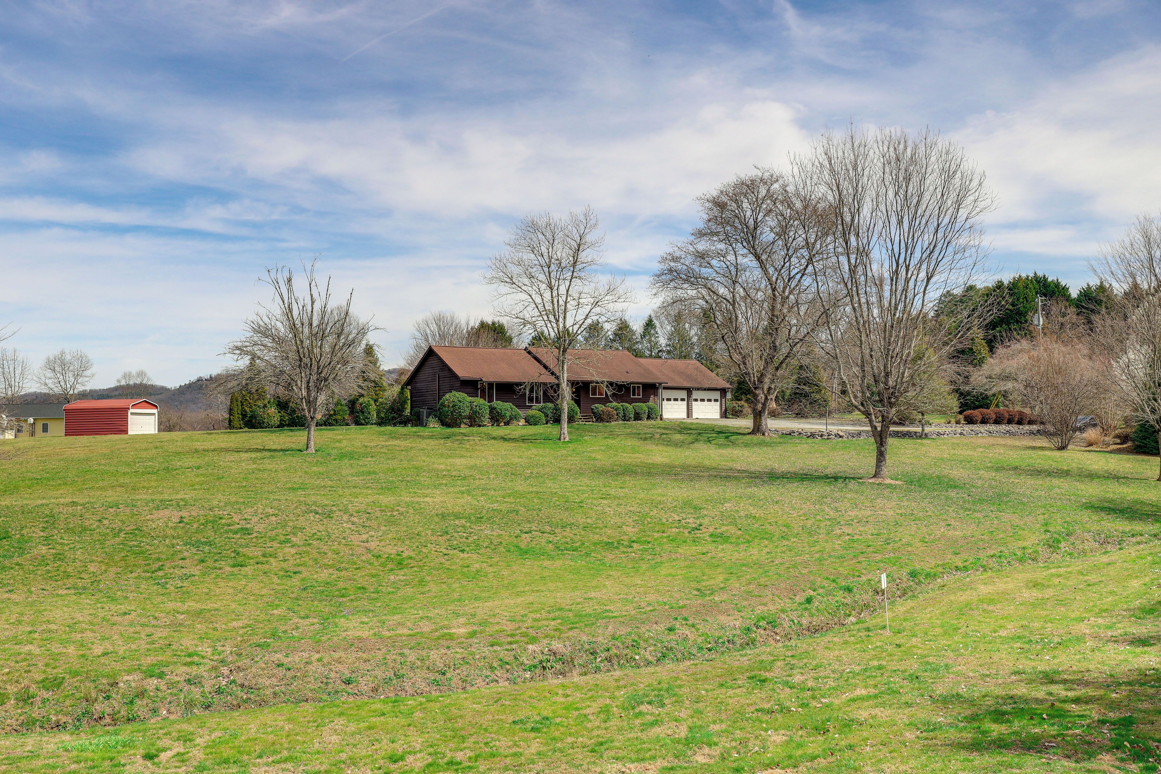 A rural property with a large open grassy field, a wooden house with a red roof, and a small shed.