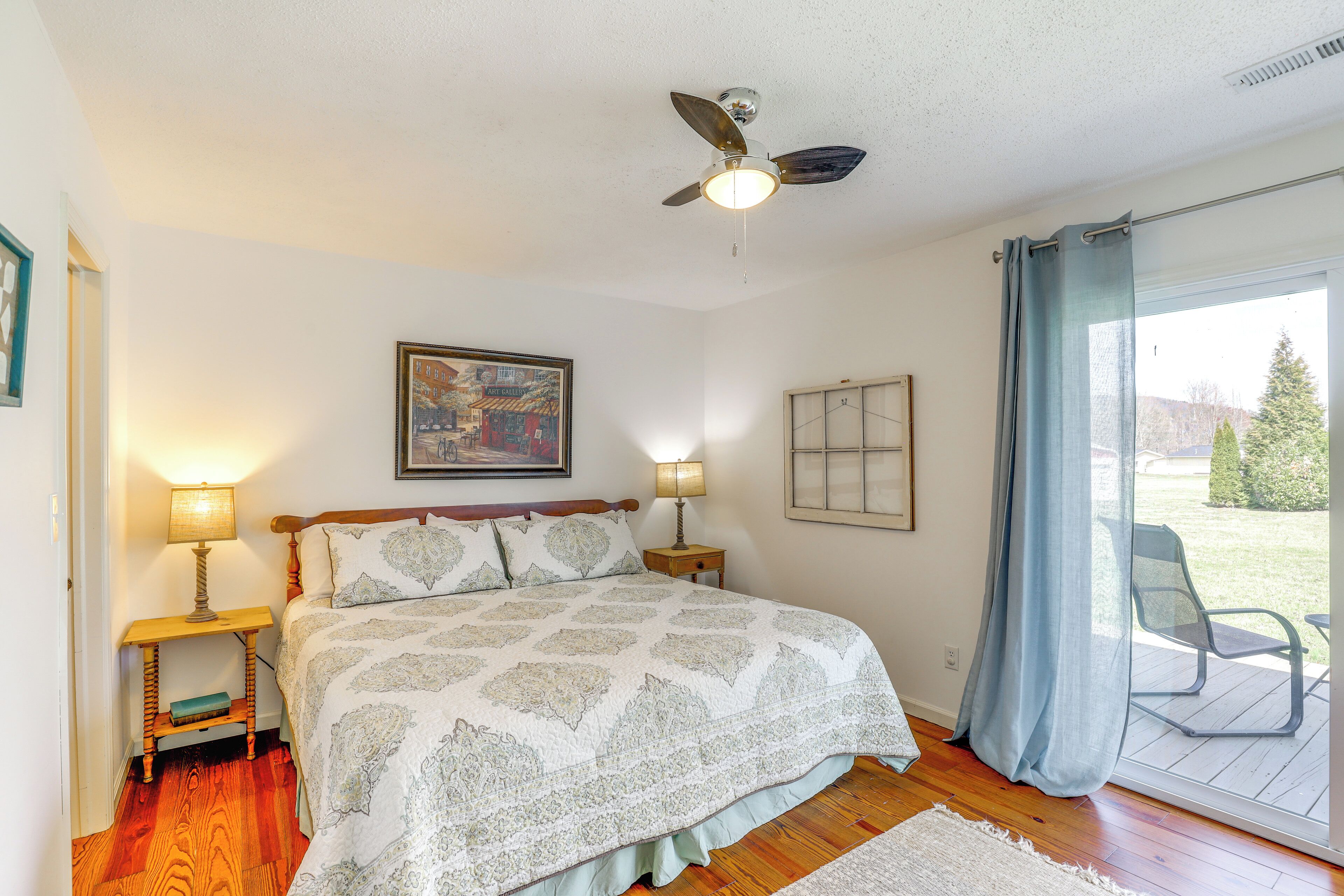 Quarto com cama, duas luminárias de cabeceira, ventilador de teto, um quadro e vista pela janela com cortina.