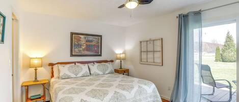 Quarto com cama, duas luminárias de cabeceira, ventilador de teto, um quadro e vista pela janela com cortina.