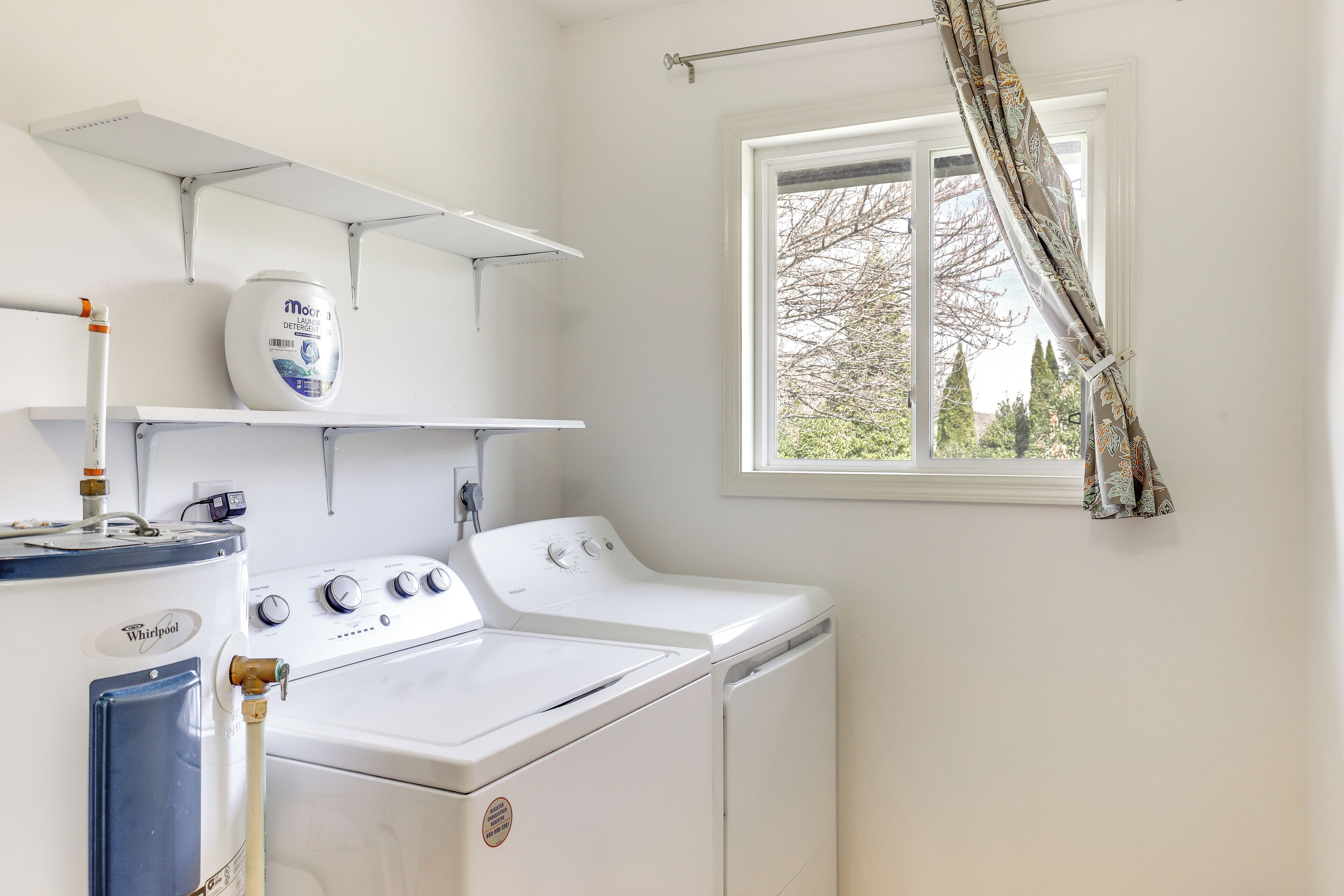 A laundry room with a Whirlpool washer and dryer, a white shelf with a detergent container, and a window with a curtain.