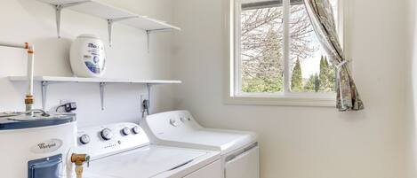 A laundry room with a Whirlpool washer and dryer, a white shelf with a detergent container, and a window with a curtain.