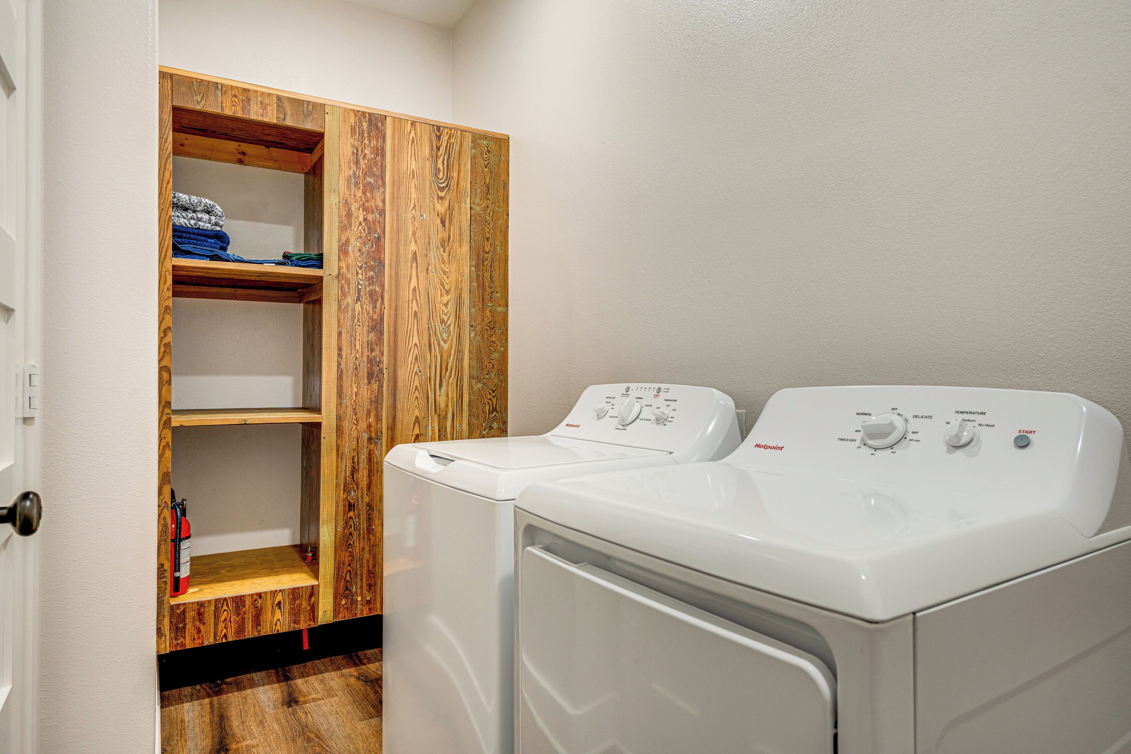 Une buanderie avec deux machines à laver et deux sèche-linge superposés, une étagère en bois où sont pliés les vêtements, et une porte sur le côté gauche.