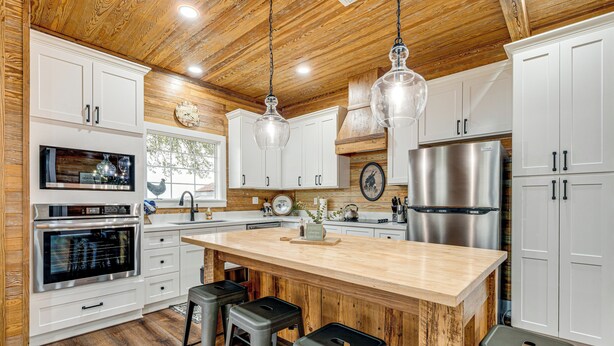 A modern kitchen with white cabinetry, a large island with stools, stainless steel appliances, and wooden ceiling beams. - Furnished Deck + Views: Rural Schulenburg Retreat (Schulenburg)