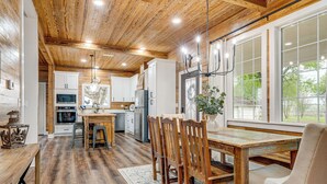 A rustic kitchen with wooden floors, a dining area with a wooden table and chairs, and a large window with a view of greenery. - Furnished Deck + Views: Rural Schulenburg Retreat (Schulenburg)
