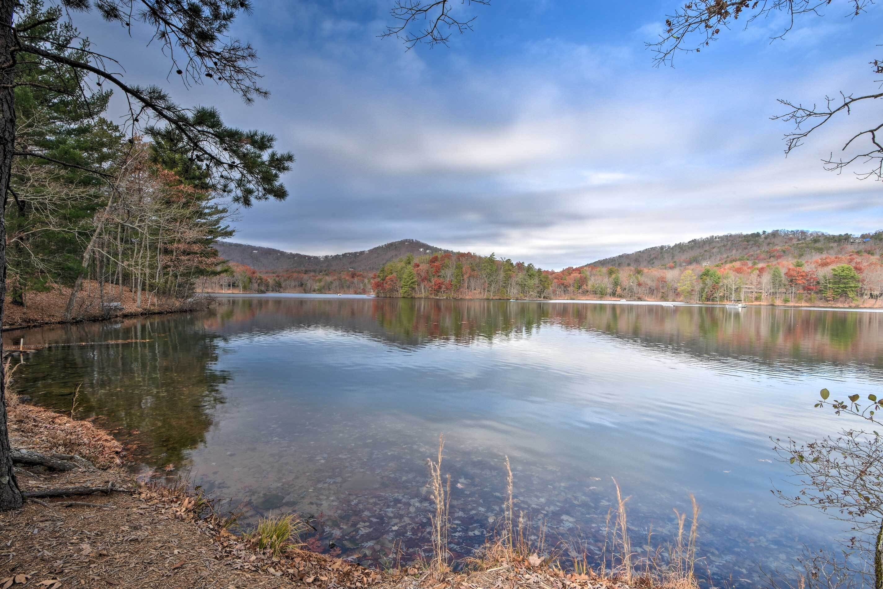 A calm lake surrounded by trees with autumn foliage, a clear sky, and distant hills.