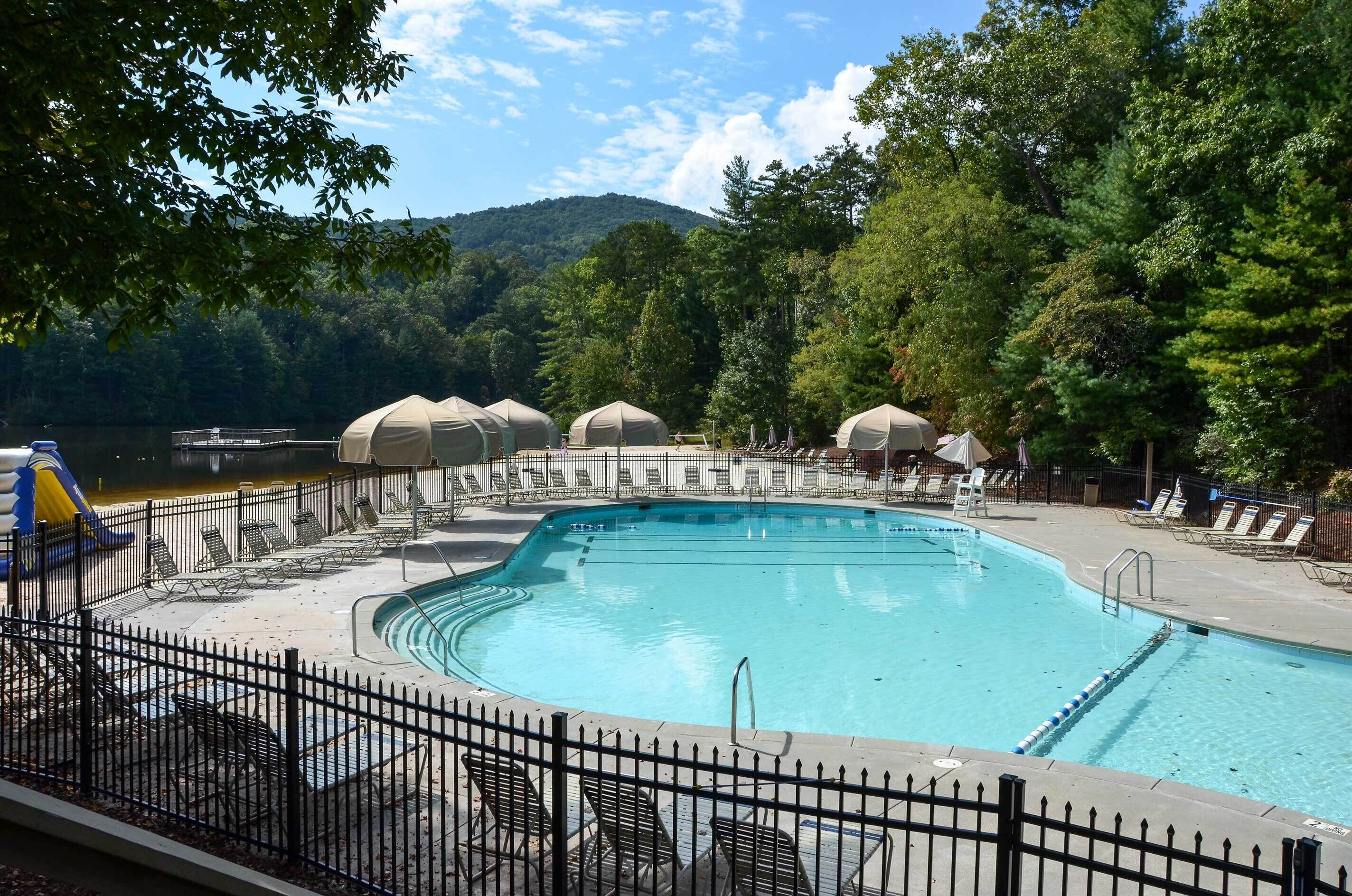 A swimming pool with lounge chairs and umbrellas, surrounded by trees and a view of a lake. - Serenity Now: Big Canoe Mountain Home! (Jasper)