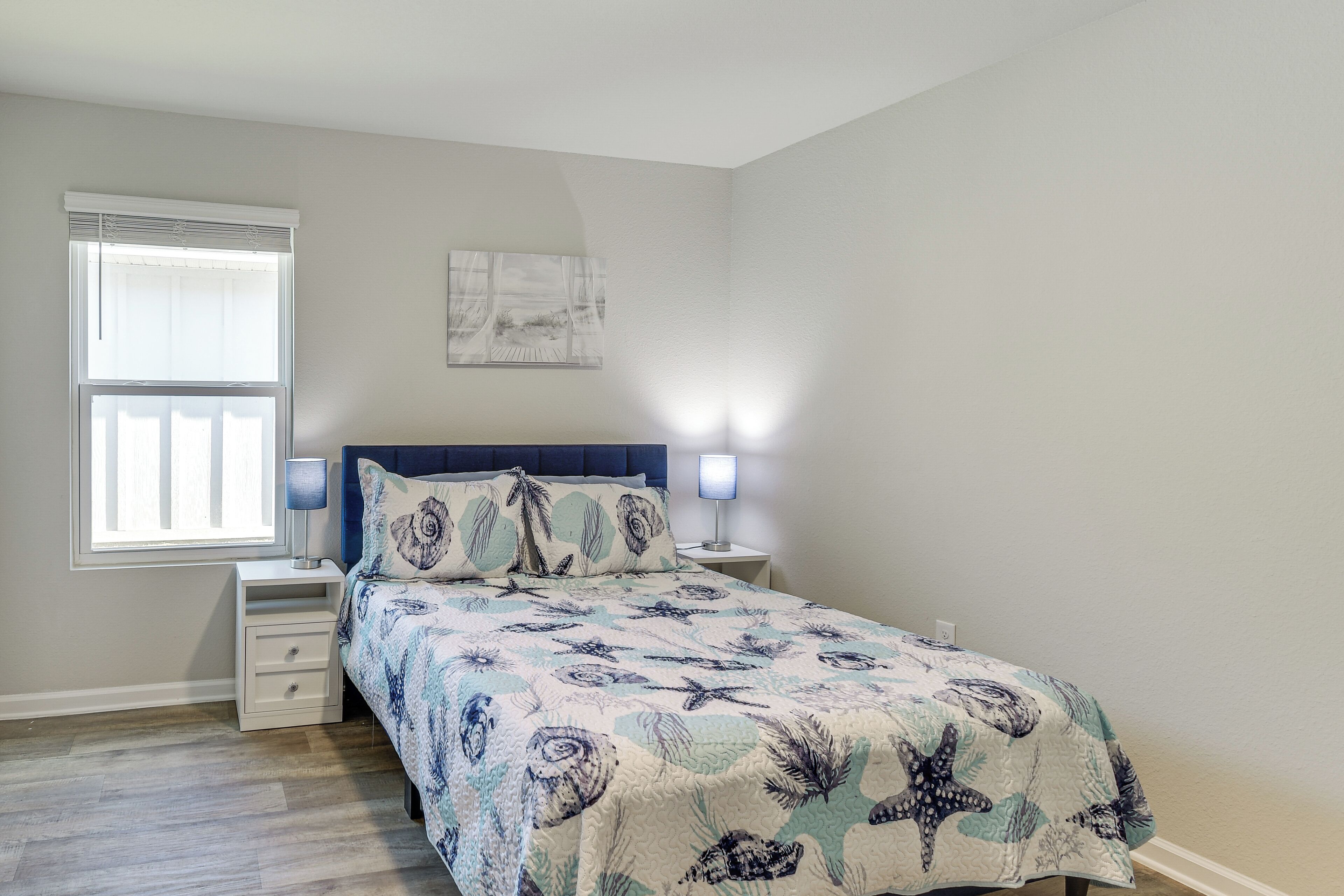 A bedroom with a bed, nightstands, a window with blinds, and a wall-mounted light fixture.
