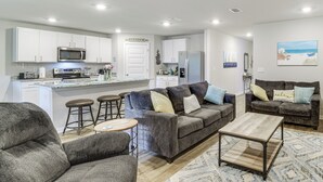 Un salon moderne avec un canapé gris, une table basse et un coin cuisine doté de meubles blancs et d’appareils électroménagers.