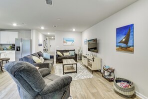 A modern living room with a sofa, coffee table, and a TV. There's a kitchen area with cabinets and a refrigerator. - 6 Mi to Gulf Shores Beach: Family Getaway! (Foley)