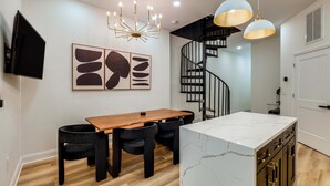 A modern kitchen with a wooden dining table, black chairs, a marble island, pendant lights, and a black spiral staircase.