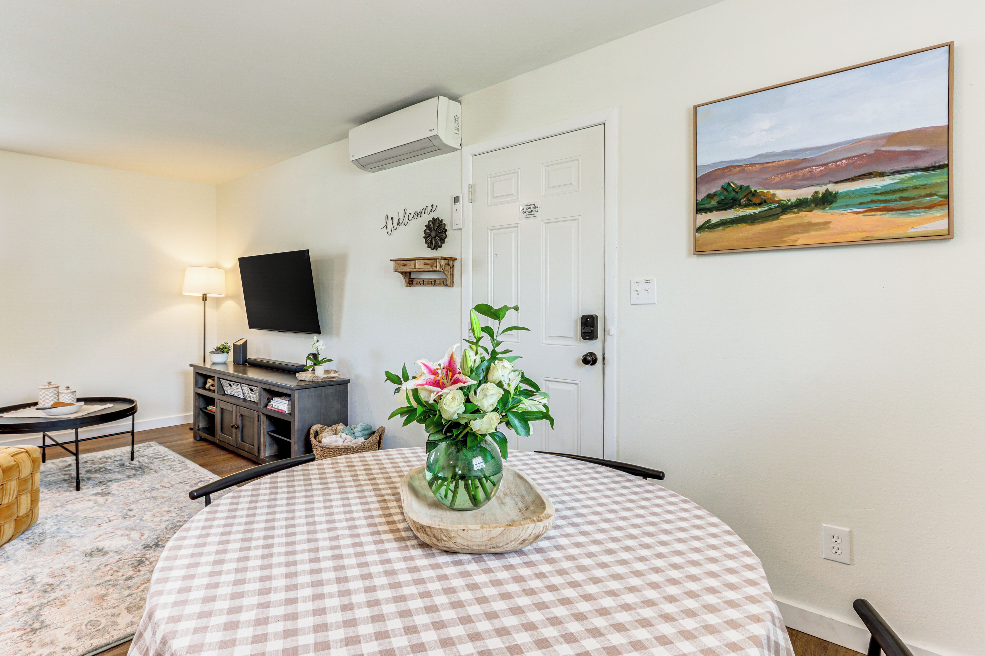 Una habitación con mantel a cuadros, un jarrón con flores, un televisor de pantalla plana, una lámpara y un cuadro en la pared.