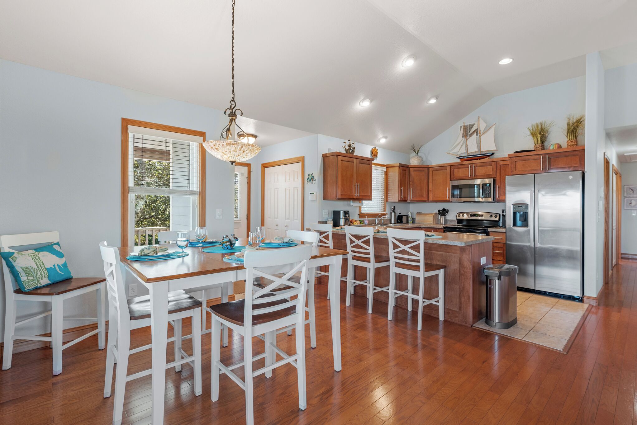 A modern kitchen with wooden cabinets, stainless steel appliances, and a dining area with white chairs and a wooden table.