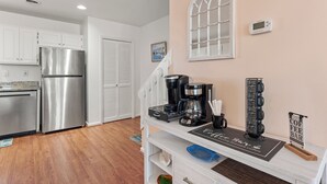 A kitchen with white cabinets, a stainless steel refrigerator, a coffee station with a coffee maker and cups, and a sign that reads "Coffee Bar." - Sea Escape: Semi-Oceanfront | Easy Beach Access (Kill Devil Hills)