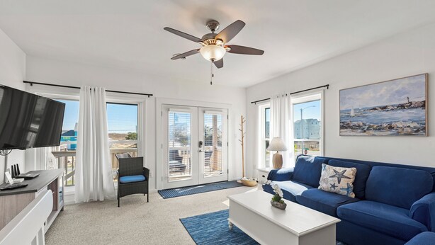 A modern living room with a ceiling fan, a white coffee table, a blue sofa, and a television on a stand. - Sea Escape: Semi-Oceanfront | Easy Beach Access (Kill Devil Hills)