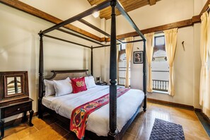 A four-poster bed with a wooden frame, white bedding, and a red and gold throw blanket. There is a nightstand with a framed picture beside the bed. The room has wooden beams on the ceiling and a window with curtains.