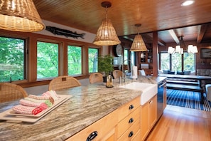 A kitchen with a granite countertop, wooden cabinets, wicker pendant lights, and a large window overlooking greenery.