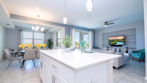 A modern kitchen with a central island, dining area with chairs, a sofa, and a TV mounted on the wall.