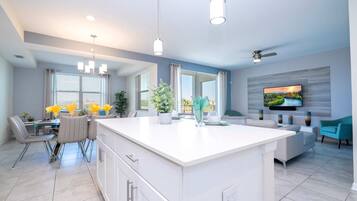 A modern kitchen with a central island, dining area with chairs, a sofa, and a TV mounted on the wall.