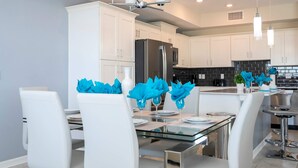 Una cocina moderna con armarios blancos, un refrigerador de acero inoxidable y un comedor con sillas blancas y una mesa de vidrio.