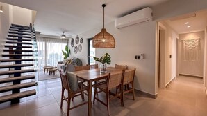 A modern living room with a dining area, featuring a wooden dining table, wicker chairs, and a hanging light fixture. - 4 Bedroom Beach Penthouse w Private Rooftop (Punta Cana)