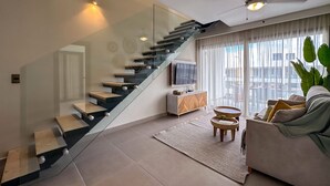 A modern living room with a glass staircase, a flat-screen TV, a sofa, and a large window with curtains. - 4 Bedroom Beach Penthouse w Private Rooftop (Punta Cana)