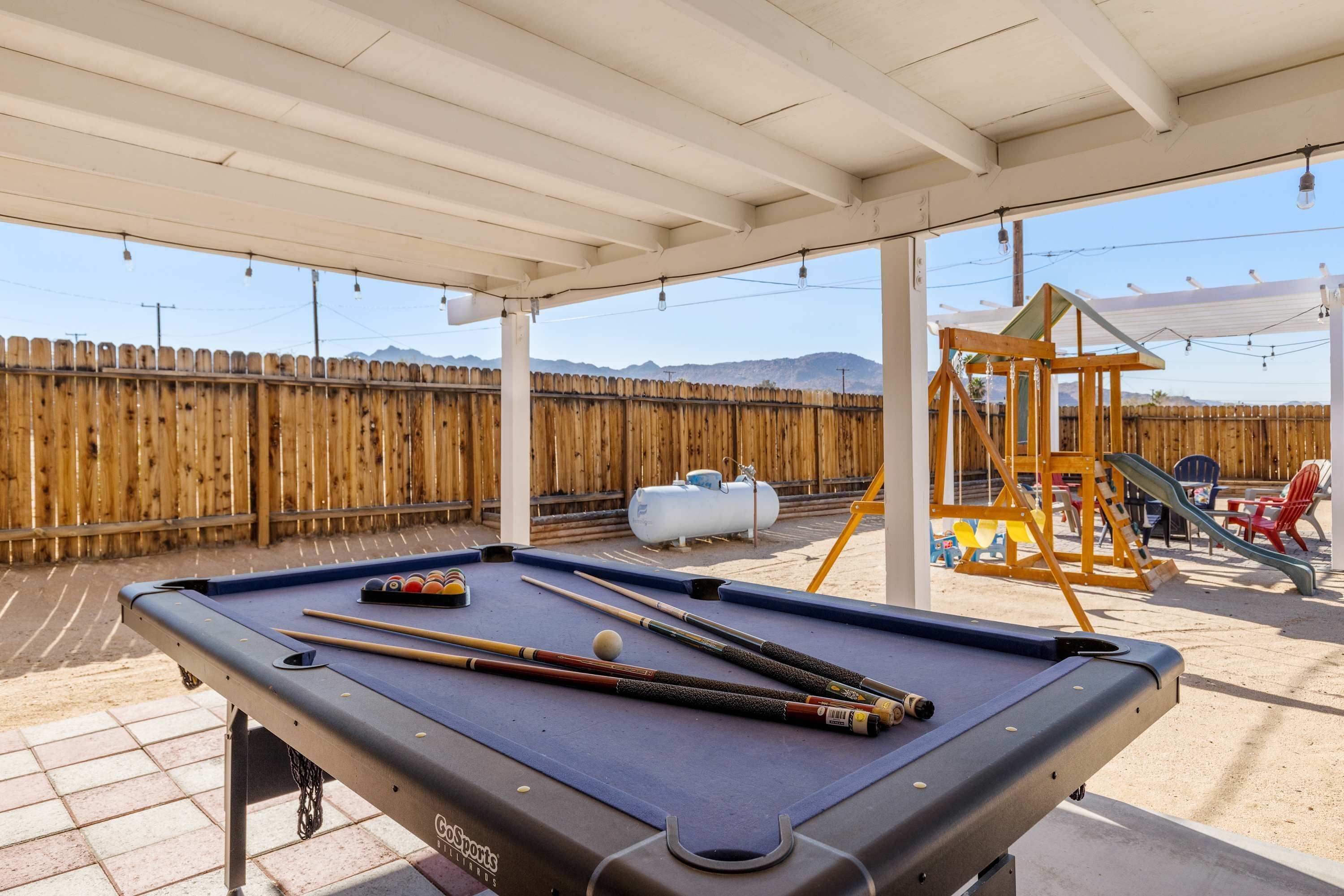 Une table de billard avec des queues et des billes, une clôture en bois et, au fond, une structure de jeux pour enfants.