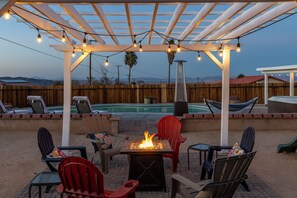 Un jardin avec un foyer, un espace salon extérieur, une piscine et une pergola illuminée de guirlandes lumineuses.
