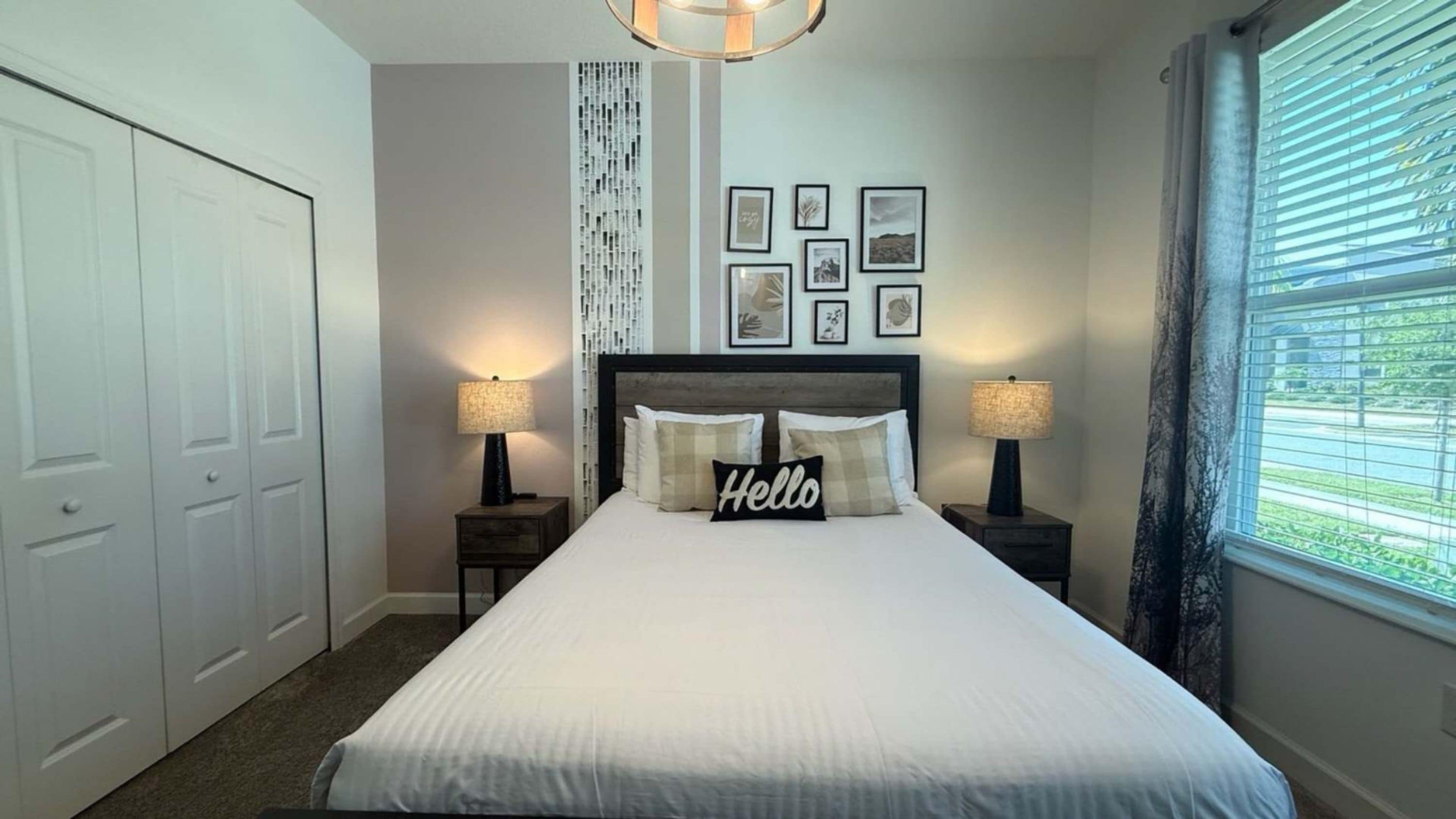 Una camera da letto ordinata con un letto grande, due comodini con lampade, una testiera con un cuscino con la scritta 'Hello', una finestra con persiane e una parete con quadri.