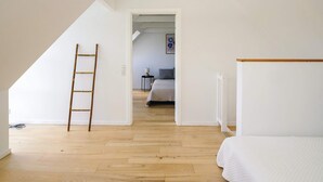 A room with a wooden floor, a bed, a ladder, and a doorway leading to another room with a bed and a lamp. - 5 Star Holiday Home in Liseleje (Liseleje)
