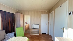 A compact room with a bed, a green chair, a small kitchenette, and a white door. - Cozy bungalow vacation idyll (Koserow)
