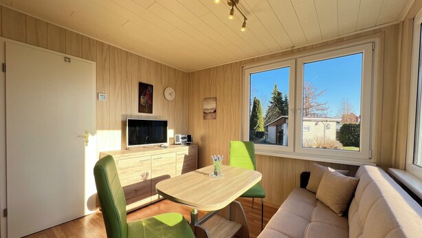 A cozy living room with a wooden floor, a sofa, a dining table with chairs, a television, and a window with a view of trees and buildings. - Cozy bungalow vacation idyll (Koserow)