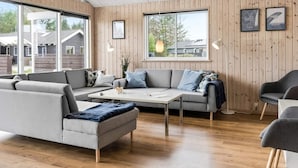 A modern living room with a wooden paneled wall, a grey sofa, a coffee table, and a view of the outside. - 5 Star Holiday Home in Albaek (Aalbaek)