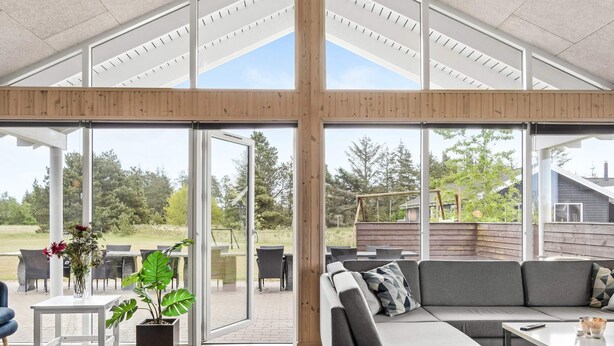 A spacious living area with a large sectional sofa, a coffee table, and a dining table. There is a potted plant and a small table with chairs. The room has large glass doors leading to an outdoor patio area with a wooden deck and outdoor furniture. - 5 Star Holiday Home in Albaek (Aalbaek)