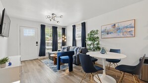 Un salon moderne comprenant un canapé, une table à manger avec des chaises, un téléviseur fixé au mur et une grande fenêtre avec des rideaux.