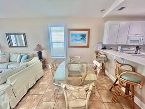 Una sala de estar con una mesa de comedor de cristal rodeada de sillas de mimbre, un sofá color crema y una ventana con persianas.