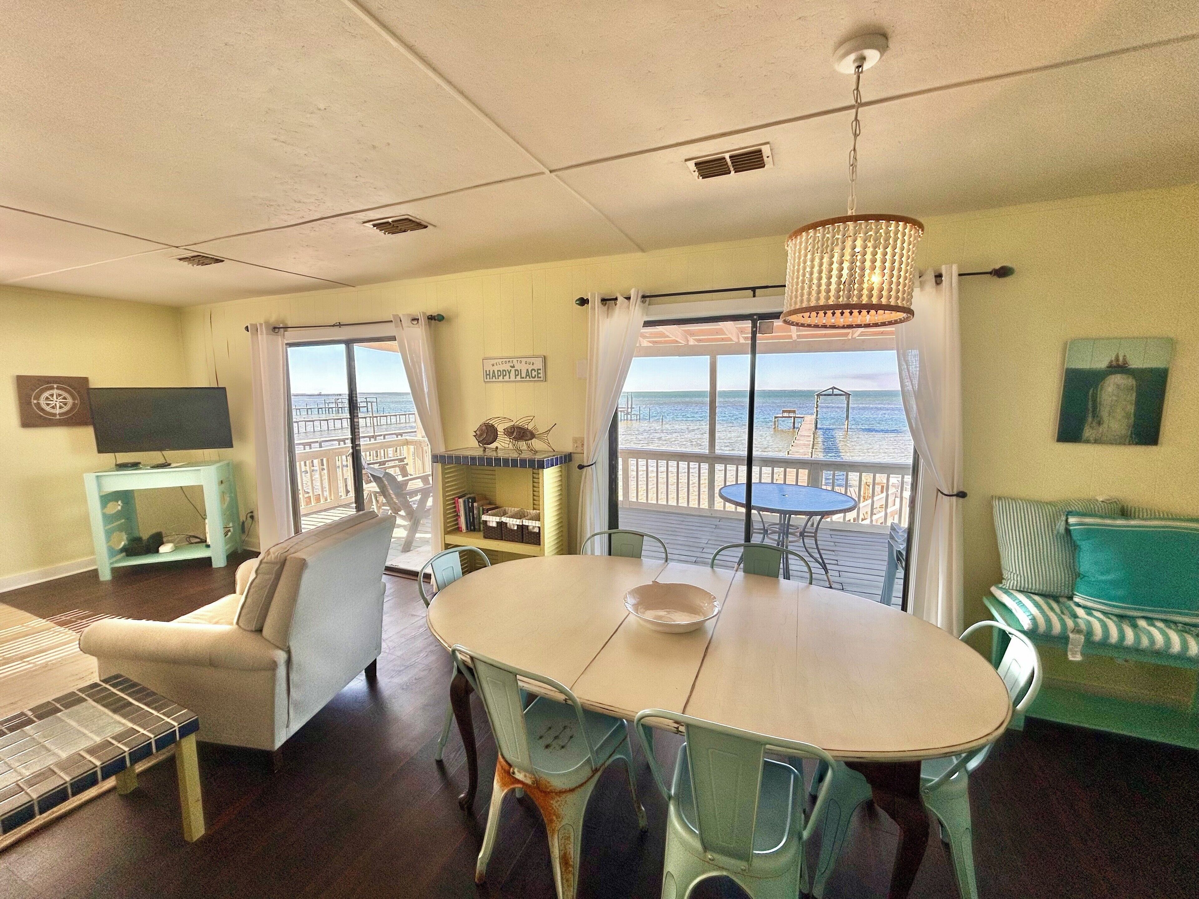 A room with a dining table, chairs, a sofa, a TV on a stand, and a view of the ocean through large windows.