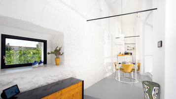 A modern interior with a wooden bench, a dining area with yellow chairs, a kitchen in the background, and a window with a view of greenery.