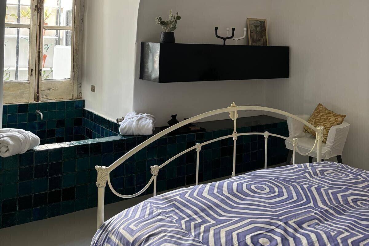 Een slaapkamer met een gebogen plafond, een bed met een beddeken met patroon, een witte stoel, een zwarte plank met decoratieve objecten en een raam met uitzicht op buiten.