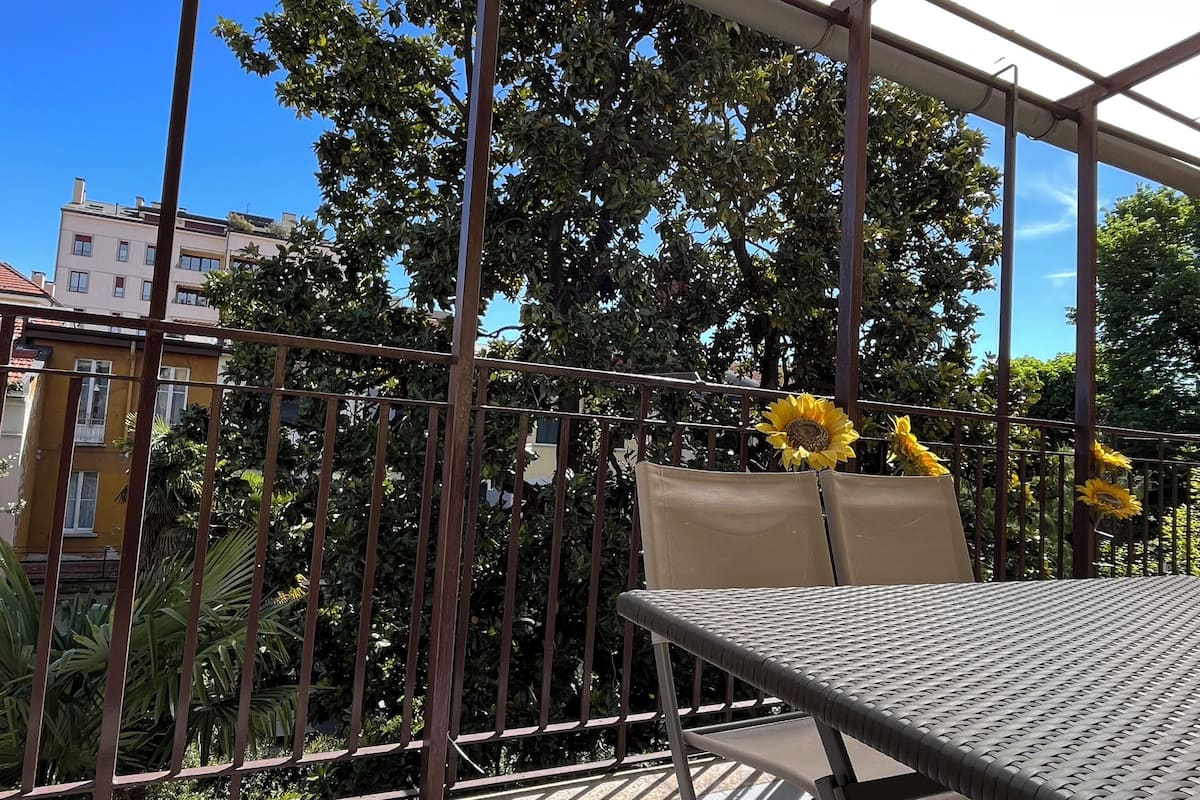 A covered outdoor seating area with a table and chairs, surrounded by a metal railing and a glass canopy.