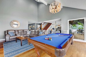 A modern living room with a pool table, a sofa, and a staircase leading to an upper level. - The Tiki House - Stay for 28! (Osage Beach)