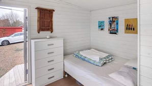 A small, single-bed room with a wooden dresser, a window with a view of a car and trees, and a door leading outside. - 4 Star Holiday Home in Stromstad (Stromstad)