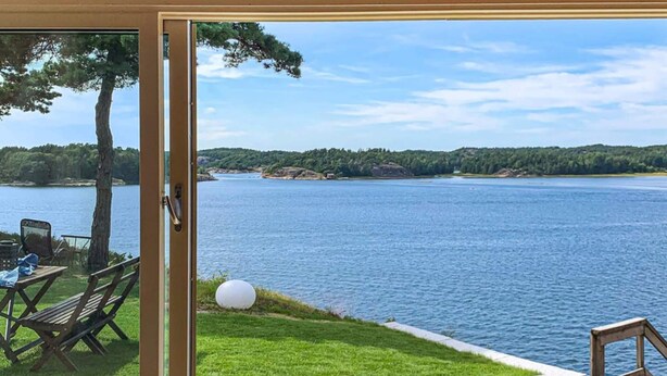A view from a room with a glass door to a deck overlooking a lake, with outdoor furniture and a pair of shoes on the deck. - 4 Star Holiday Home in Stromstad (Stromstad)