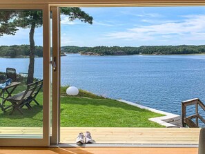 A view from a room with a glass door to a deck overlooking a lake, with outdoor furniture and a pair of shoes on the deck.