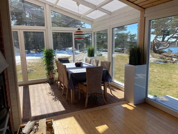 A sunlit room with a dining table and chairs, large windows, and a view of a natural landscape.