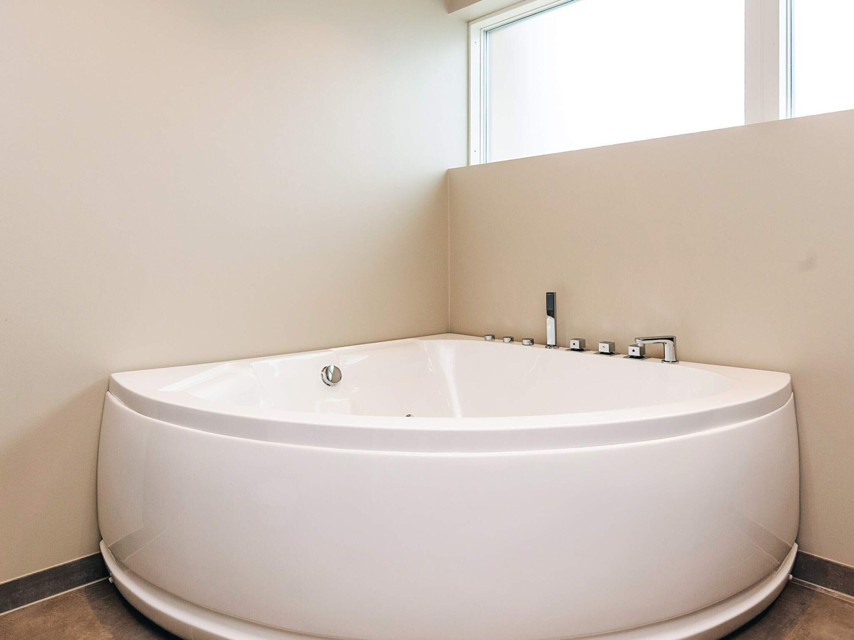 Une grande baignoire blanche, autoportante, placée dans un coin d’une pièce aux murs sobres, avec une fenêtre au-dessus.
