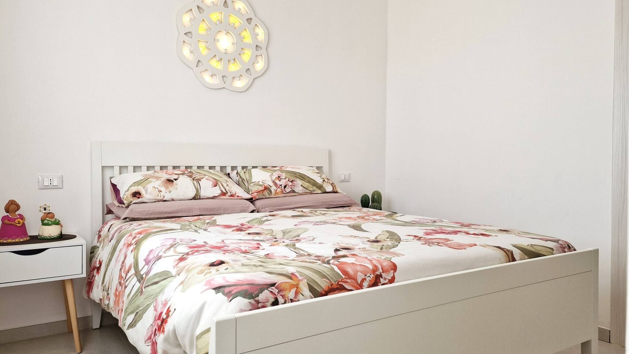 Une chambre avec un lit blanc, une literie à motifs floraux, deux tables de chevet, un luminaire fixé au mur et deux petits cactus posés sur une table de chevet.
