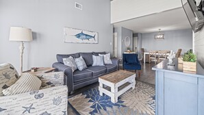 Ein modernes Wohnzimmer mit einem grauen Sofa, einem Holztisch und einem blauen Sessel. Im Hintergrund befindet sich ein Essbereich mit Tisch und Stühlen.