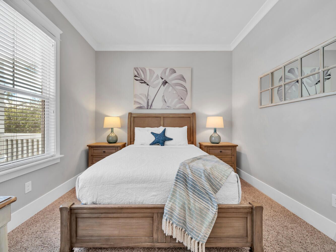 A bedroom with a wooden bed, two bedside lamps, a starfish pillow, and a large window with blinds.