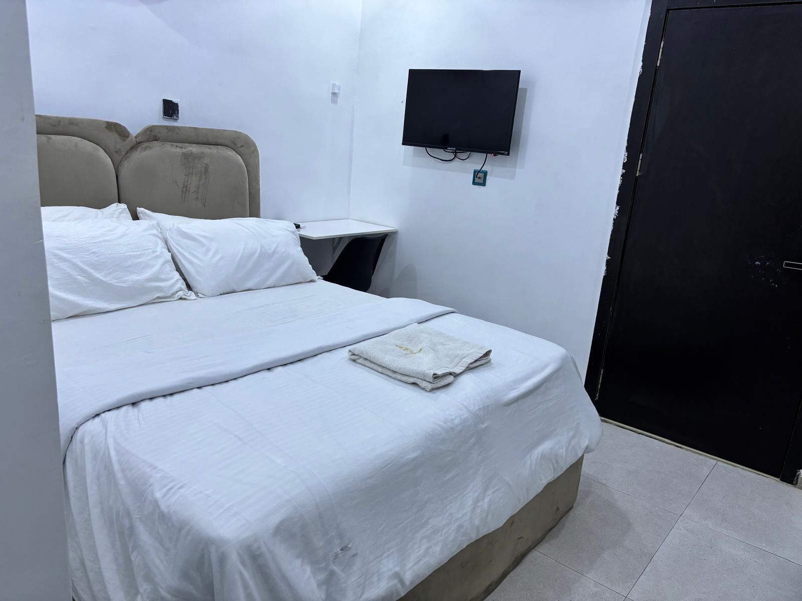 A hotel room with a bed, a TV mounted on the wall, a bedside table, and a door.